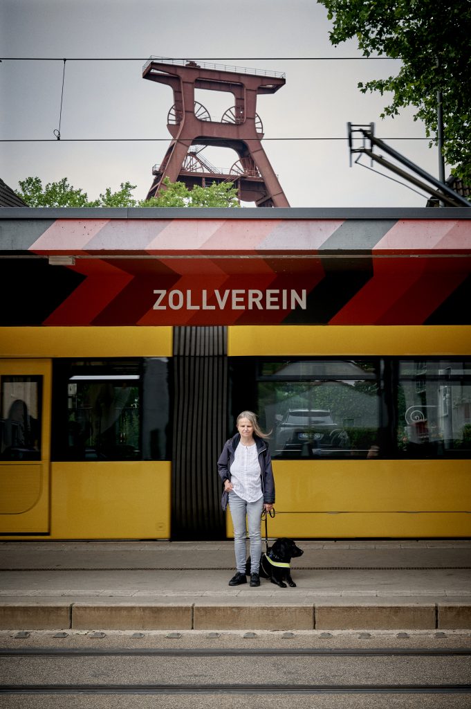 Blinde Frau mit Führhund steht auf dem Bahnsteig der Haltestelle Zollverein.