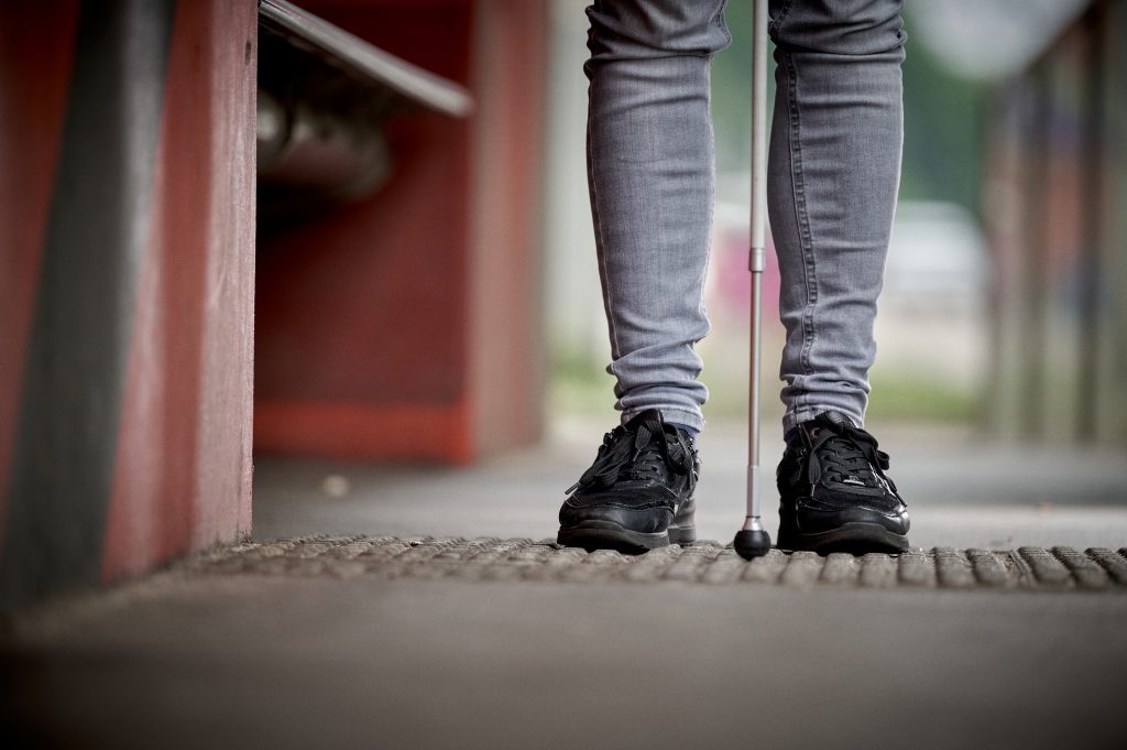 Blinde Person mit Langstock auf barrierefreiem Boden an Haltestelle.