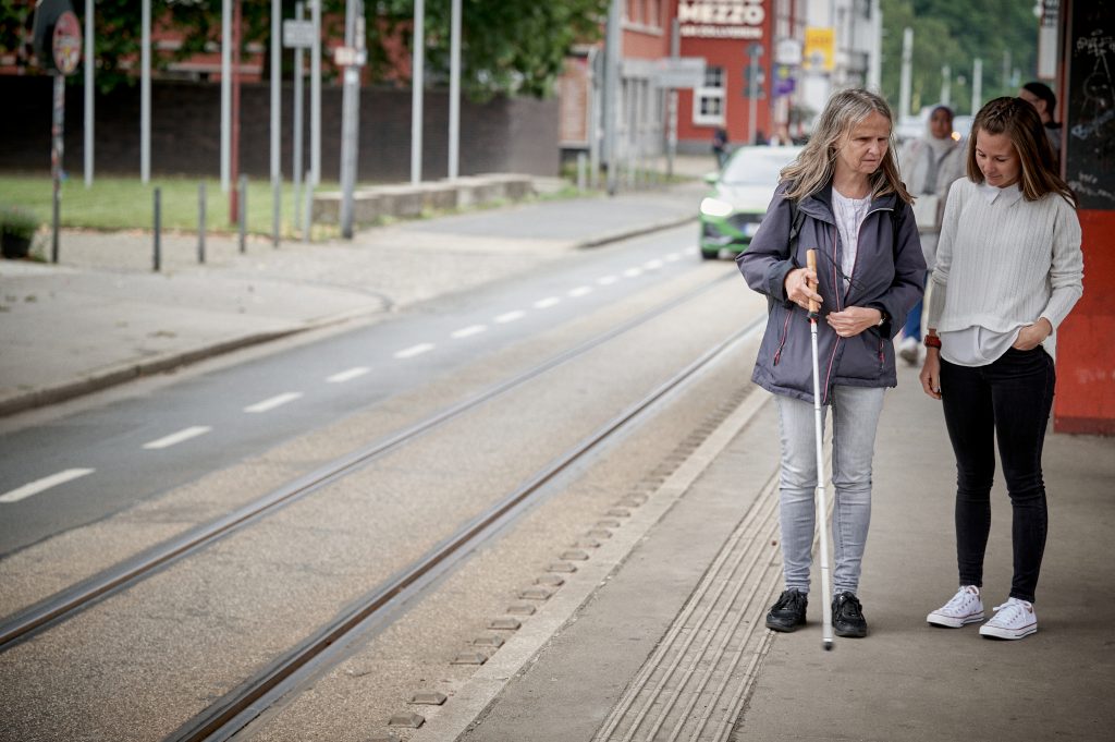 Blinde Frau mit Rehalehrerin an barrierefreier Straßenbahnhaltestelle – Mobilitätstraining und Inklusion im öffentlichen Nahverkehr.