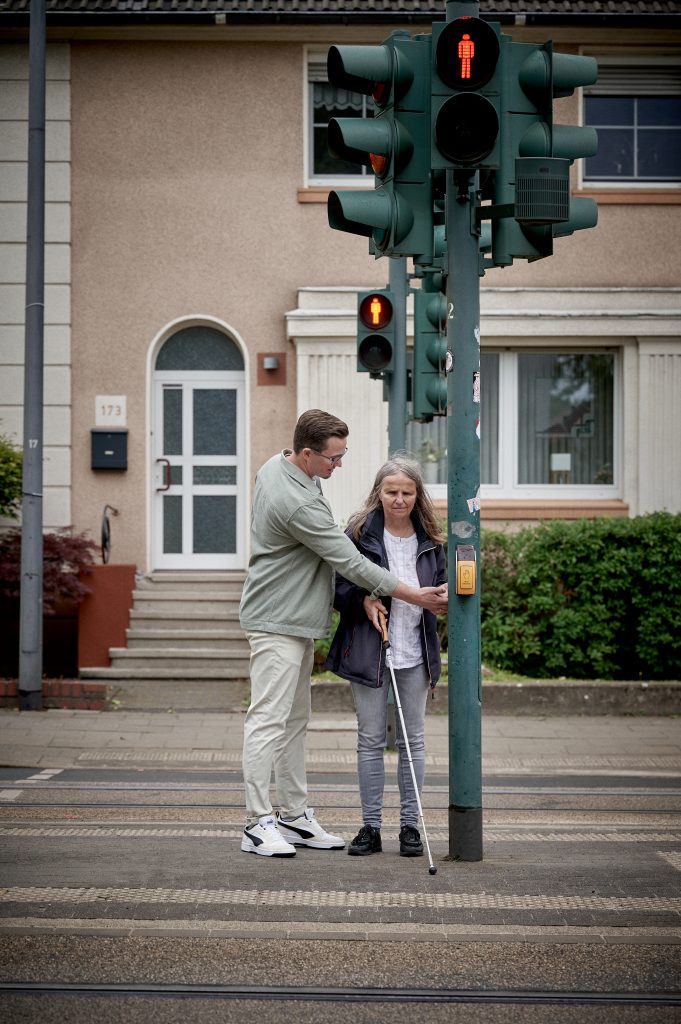 Blinde Frau mit Langstock an einer Fußgängerampel in Schulungssituation mit Rehalehrer.