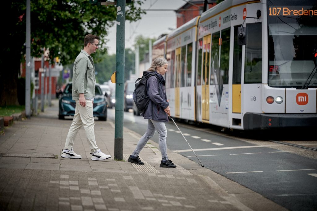 Blinde Frau mit Langstock überquert eine Straße in Schulungssituation mit Rehalehrer.
